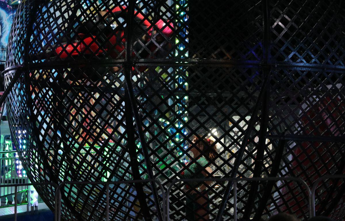 A girl stands in the middle of a sphere metal cage. A man on a motorcycle rides on the side of the cage looking down at her.