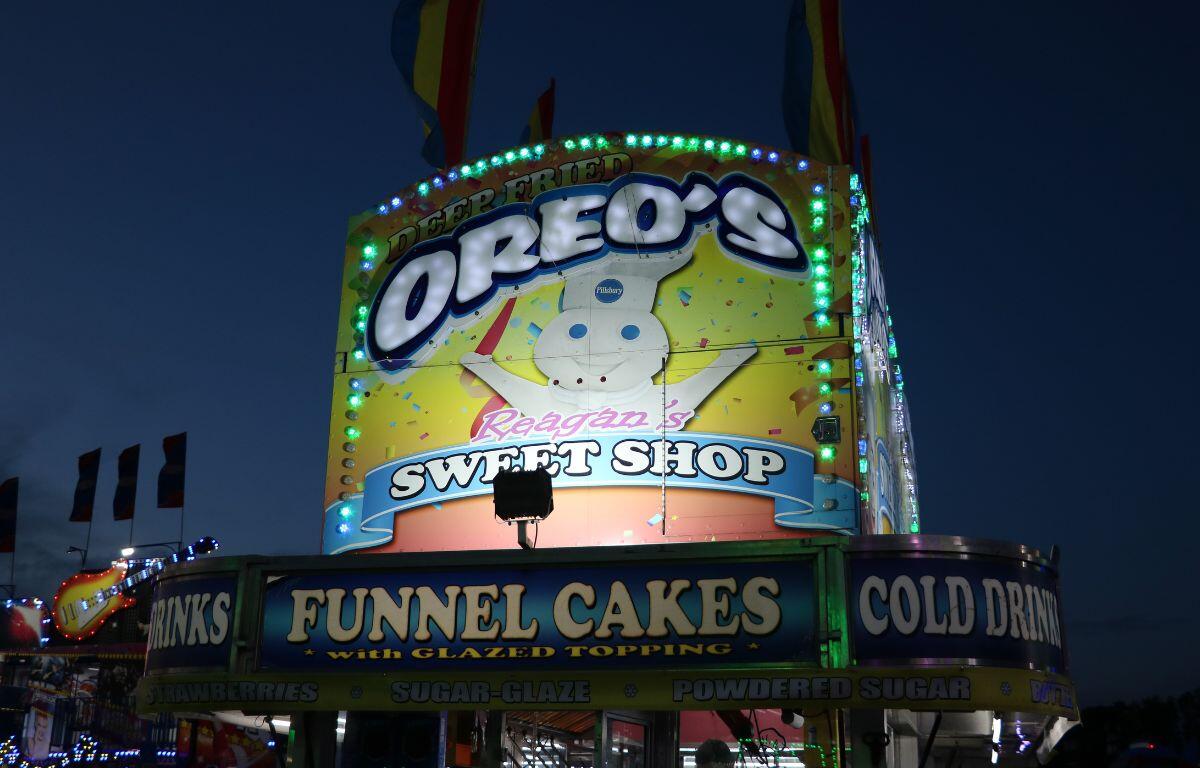 A large food stand has a light up sign at the top reading, "Oreo's. Reagan's Sweet Shop. Funnel Cakes with glazed toppings."
