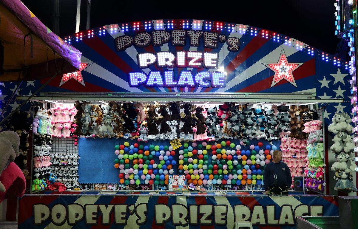 A game booth at a fair features balloons on a blue wall that guests can pop. Hanging above and around the wall are stuffed prizes. The top and bottom supports of the game read, "Popeye's Prize Palace."