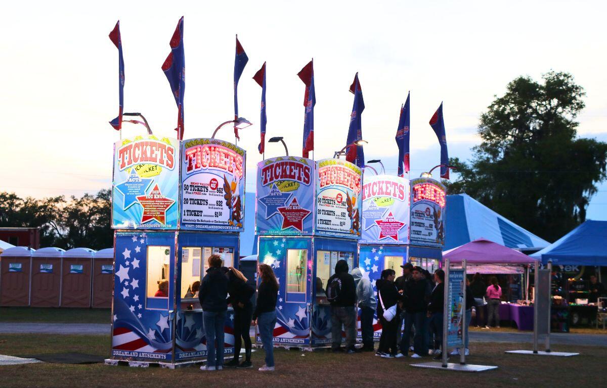 Three tickets booths are lined up in an open field at a fair with guests waiting in line for tickets.