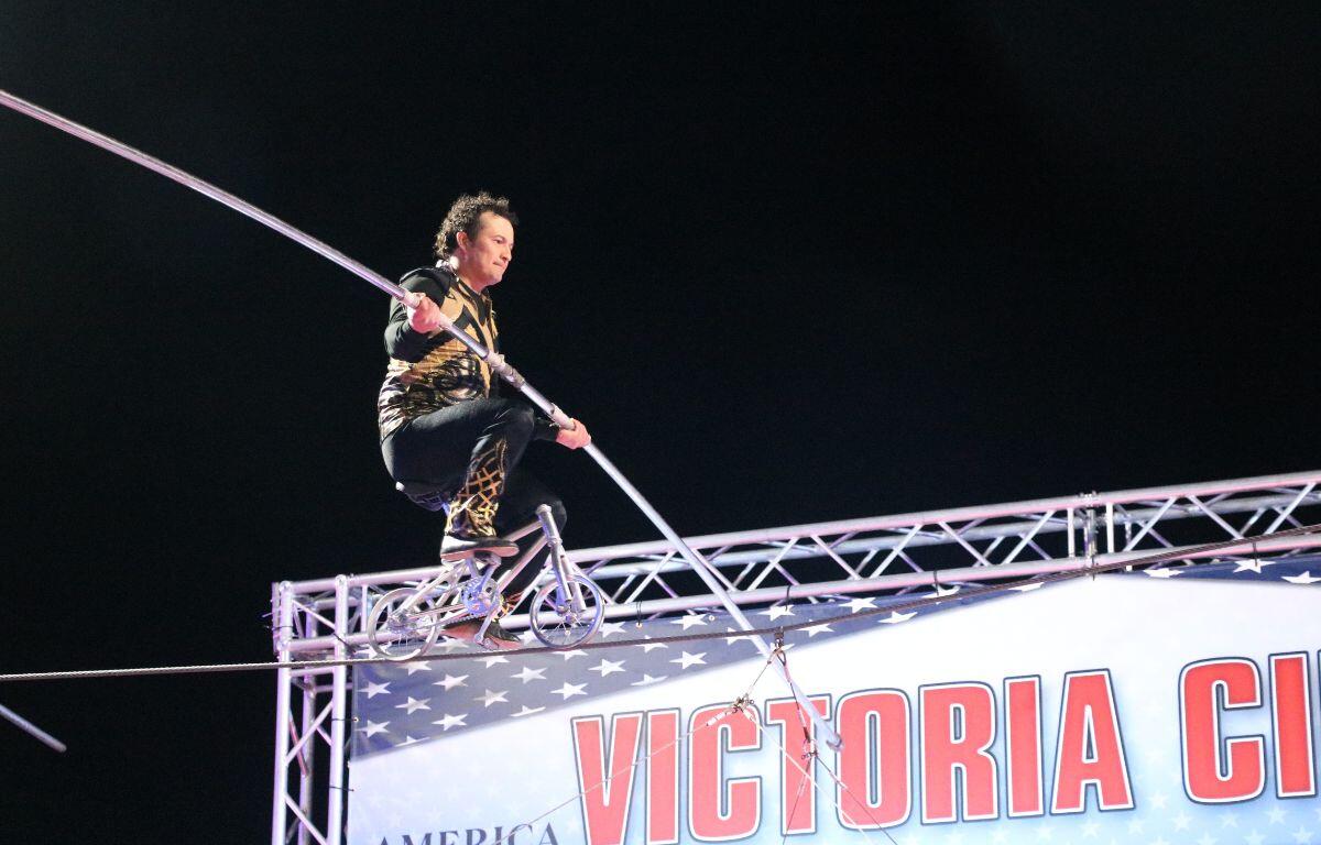 A performer rides a small bike with a large pole on a tight rope.