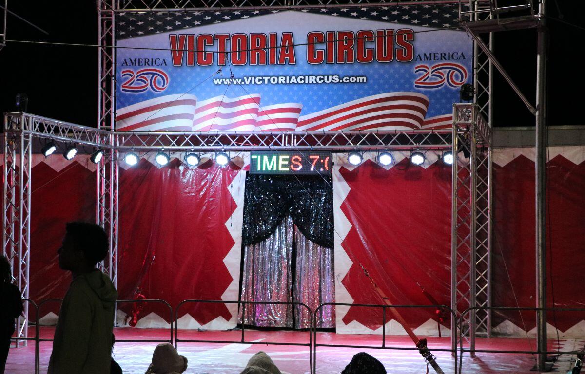 A circus performance features a large facade with red and white walls and sparkling curtains in the center. A large sign above the curtains reads, "Victoria Circus." Guests sit behind a metal barrier waiting for the show to begin.