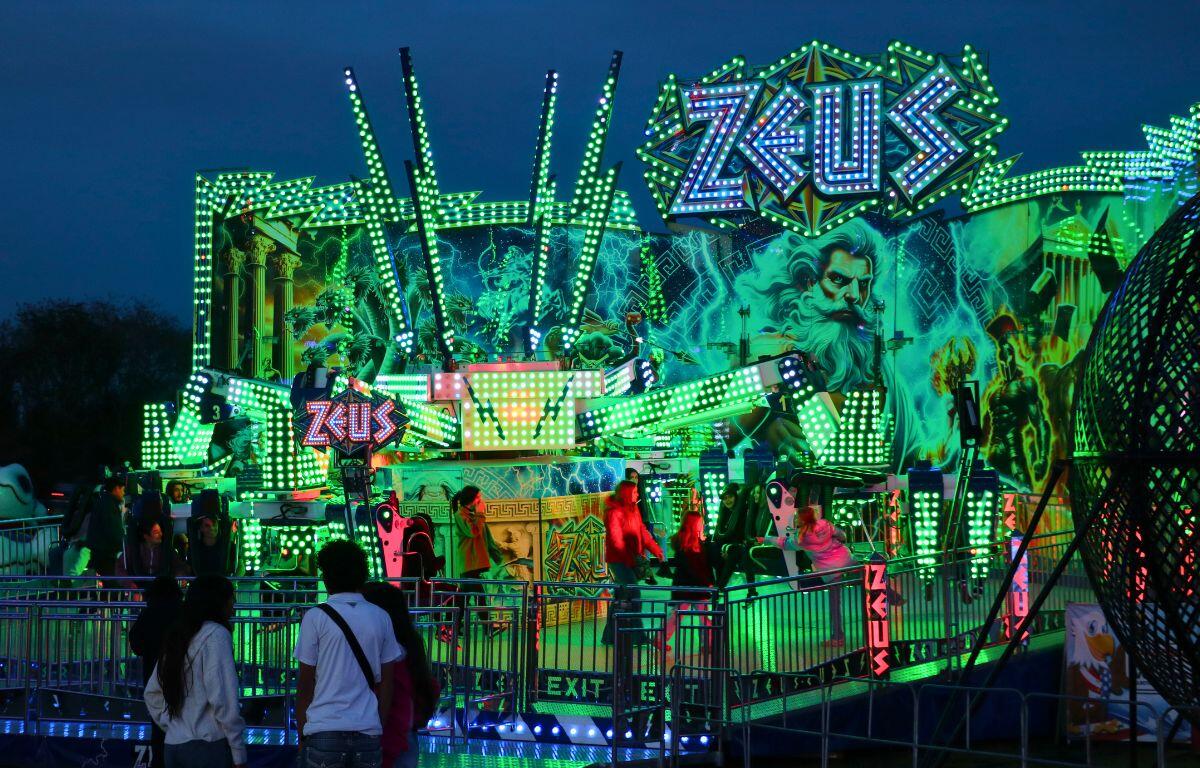 A large fair ride has seats elevated by metal supports as guests board it. The is illuminated by green lights with a large, blue sign toward the top of the backdrop facade reading, "Zues."