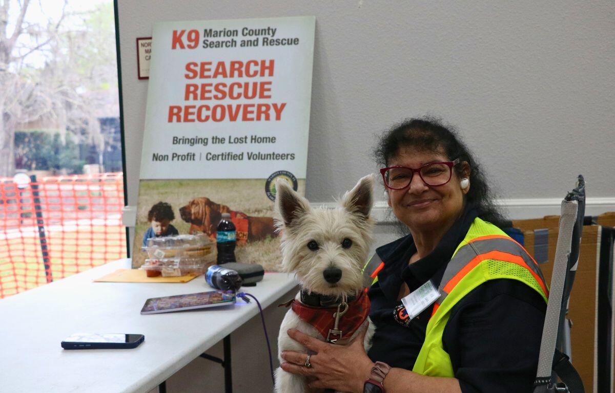 A woman and a dog sit together in front of a table with a sign in the background reading, "K9 Marion County Search and Rescue. Search. Rescue. Recovery."