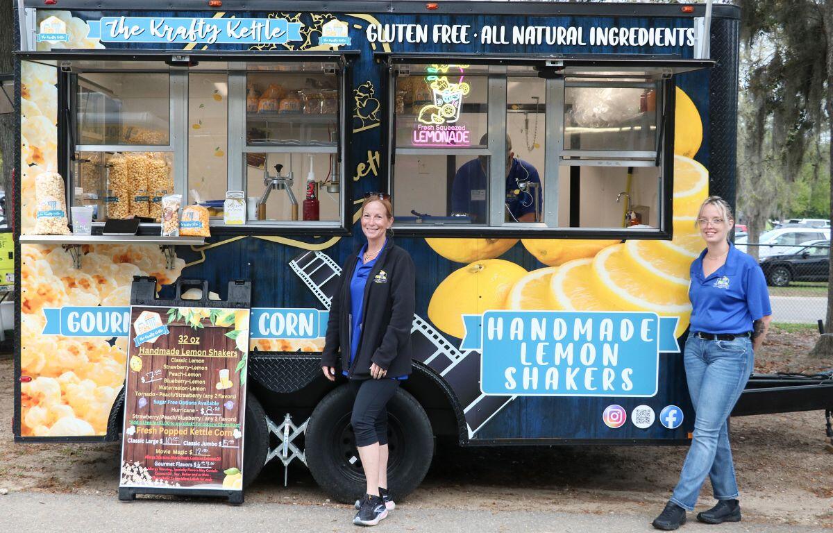 Two women stand in front of a food trucks with large lemon and popcorn designs on it. A sign in front of the truck reads a menu.