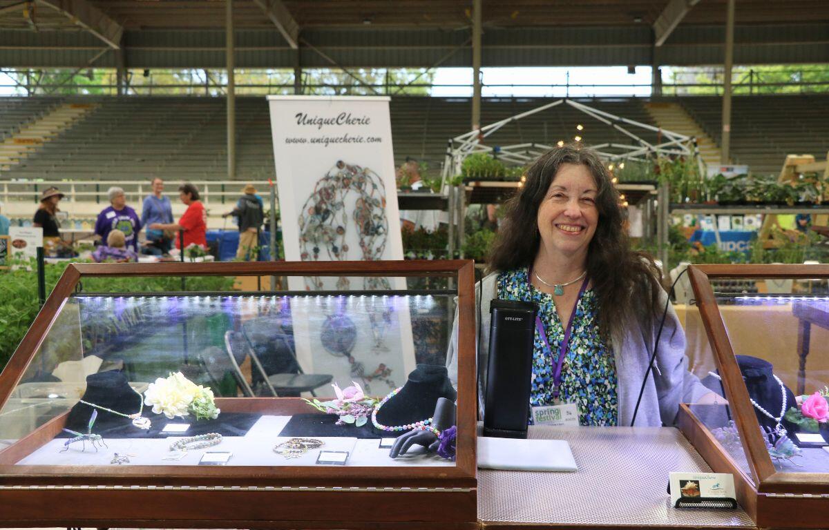 A woman stands behind display cases with flower-themed jewelry. In the background is a market garden with crowds walking around.