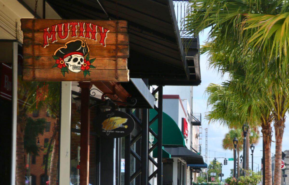 A vibrant, wooden sign reads, "Mutiny." Behind it is a line of stores on a sidewalk. On the right are palm trees.