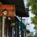 A vibrant, wooden sign reads, "Mutiny." Behind it is a line of stores on a sidewalk. On the right are palm trees.