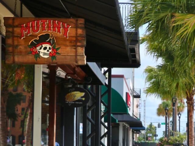 A vibrant, wooden sign reads, "Mutiny." Behind it is a line of stores on a sidewalk. On the right are palm trees.