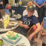 In the background, a room festooned, with garland, ornaments, adults and children. to the left tables with centerpieces and plates, in the middle of the photo a man wearing a hat sits in front of cake with candles, and to his right more adults and children.