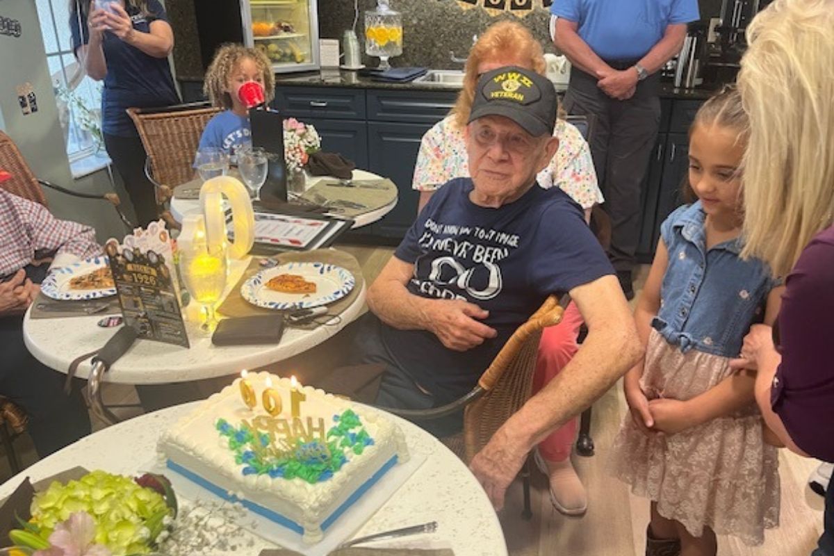 In the background, a room festooned, with garland, ornaments, adults and children. to the left tables with centerpieces and plates, in the middle of the photo a man wearing a hat sits in front of cake with candles, and to his right more adults and children.
