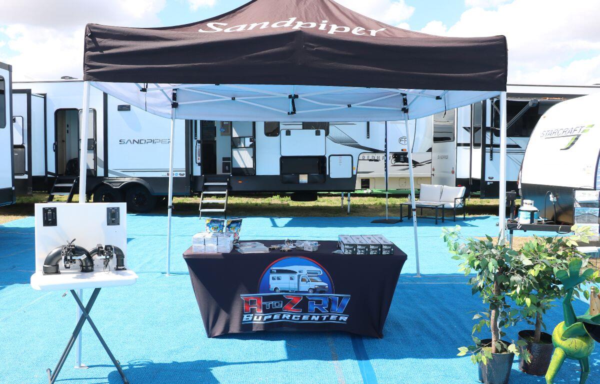 A canopy with a black top on it features white letters that reads, "Sandpiper." A table under the canopy features a black tablecloth with a logo in front of it reading, "A to Z RV Supercenter." On the table are advertised items for RVs. In the background are RVs surrounding the area.