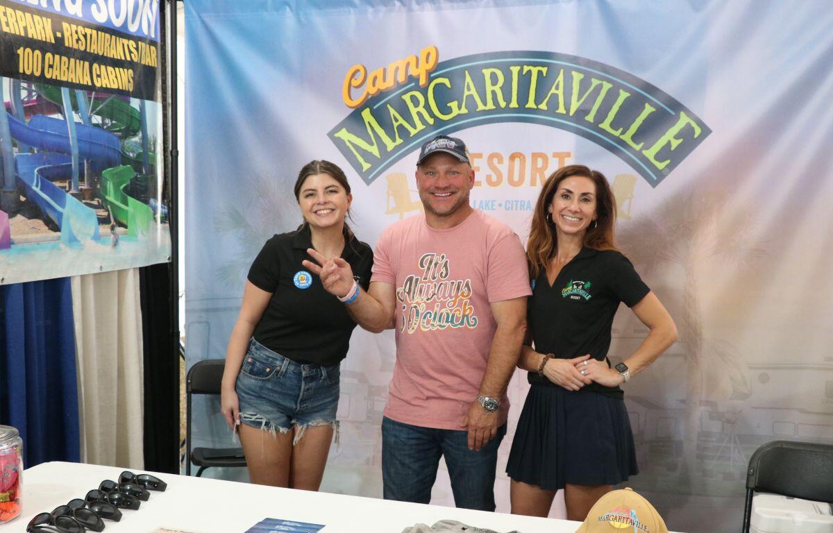 Three people stand behind a table with multiple advertising materials. In the background is a large banner reading, "Camp Margaritaville Resort."