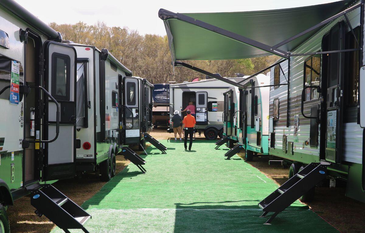 A long section of green flooring features RVs on either side in grass with the stairs out for people to enter. Toward the back are guests entering an RV.