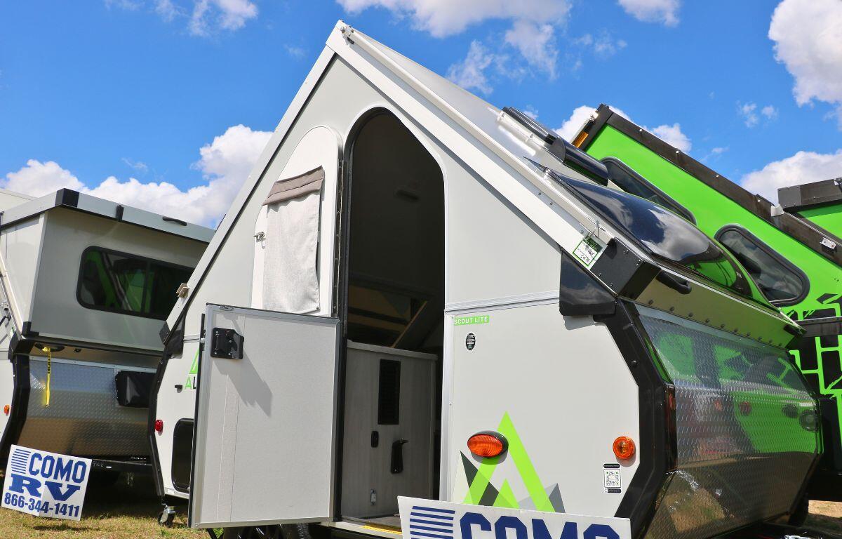 A short RV with a pointed roof features a small interior and signs reading, "Como RV. 866-344-1411." More RVs of the same design surround it.