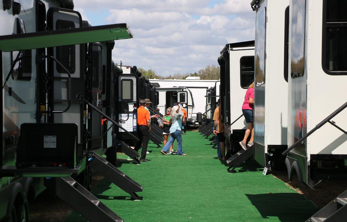 Green flooring is placed in the center of a line of multiple RVs with their stairs down. People walk around and enter the RVs.