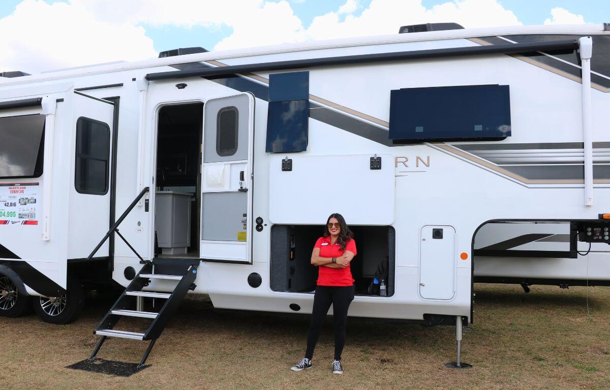 A woman in a red shirt stands in front of a large RV with the door open and stairs down. The RV stands in a grass field.