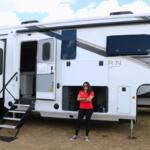 A woman in a red shirt stands in front of a large RV with the door open and stairs down. The RV stands in a grass field.