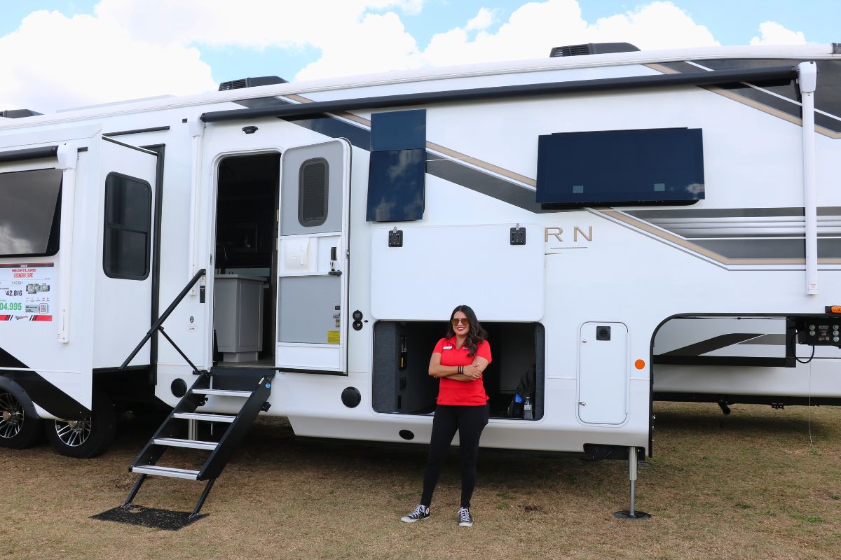 A woman in a red shirt stands in front of a large RV with the door open and stairs down. The RV stands in a grass field.