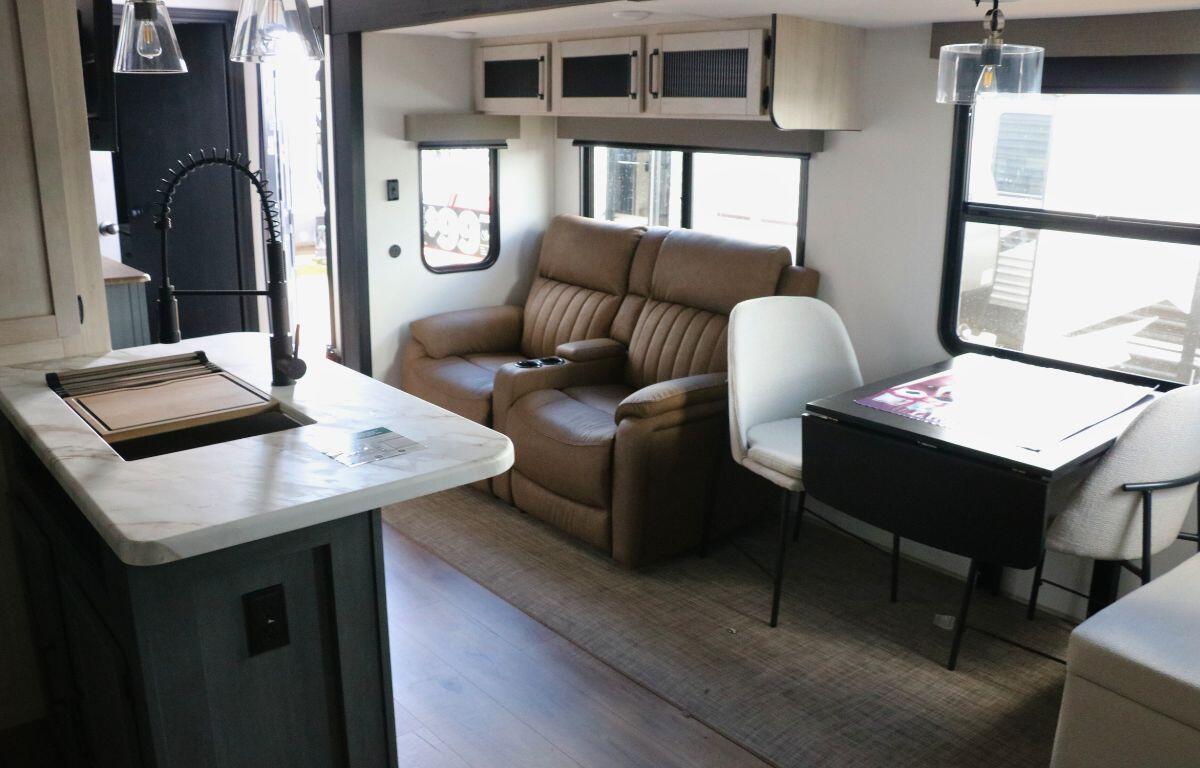 The interior of an RV features a sofa, kitchen and small dining area with large windows, hanging ceiling lights and cabinets throughout the room.
