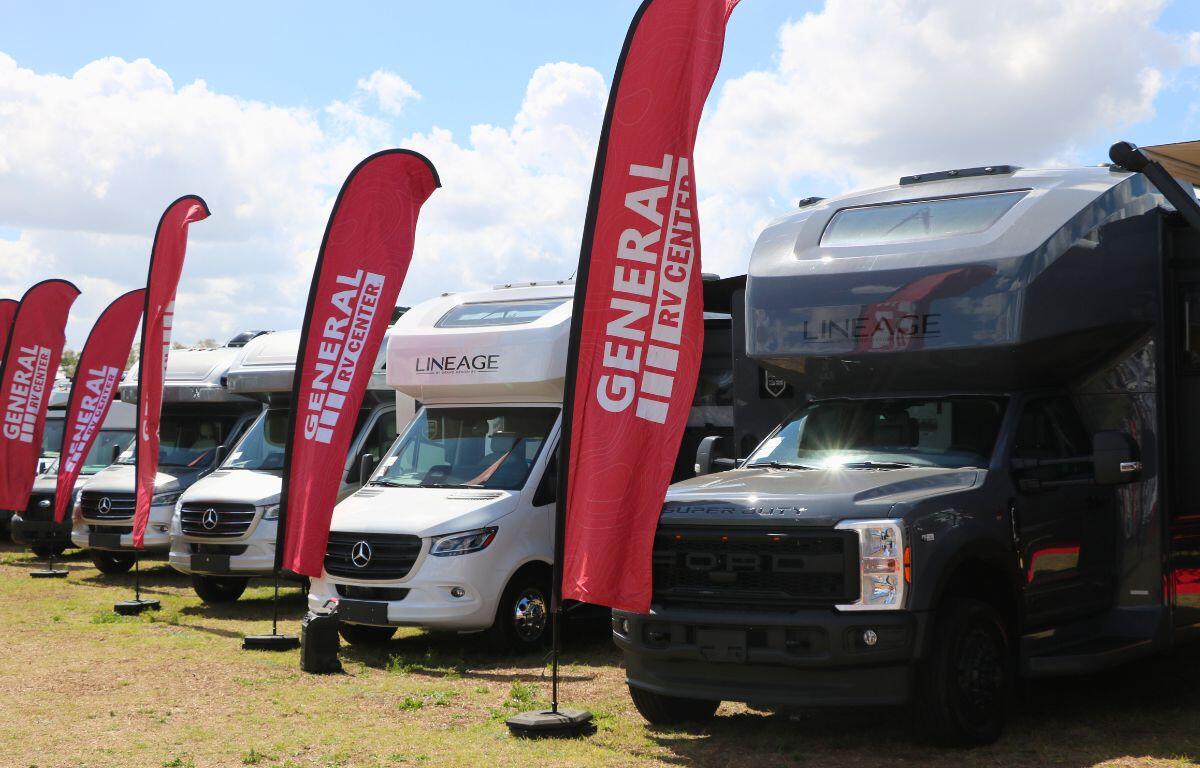RVs on trucks of varying colors are lined up in a grass field with tall, red flags placed in front of each RV. The flags have red and white letters reading, "General RV Center."