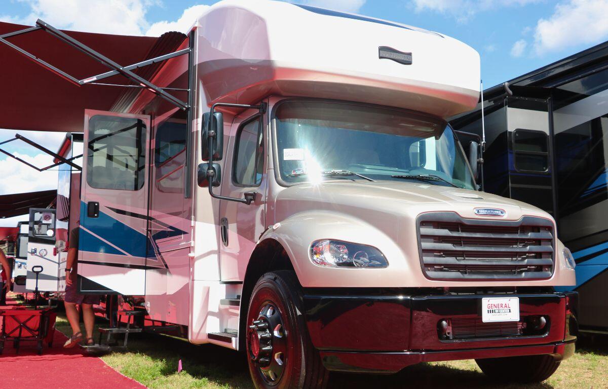 A large RV stands in a grass field with its doors open. The front plate of the RV reads, "General RV Center." In the background are more RVs and red flooring.