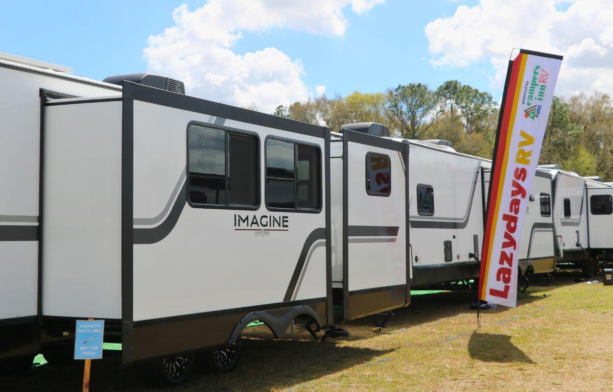 Large RVs are bumper to bumper in a grass field with a tall flag reading, "Lazydays powered by Campers Inn Rv."