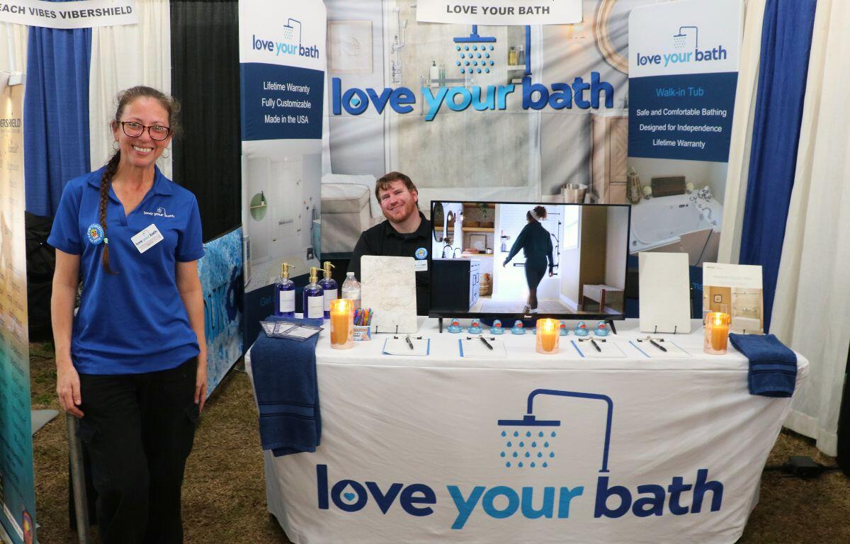 A vendor features a table with sign-up sheets, candles, ducks, a monitor and more with a white tablecloth. The tablecloth features a logo of a shower with light and navy-blue letters reading, "love your bath."