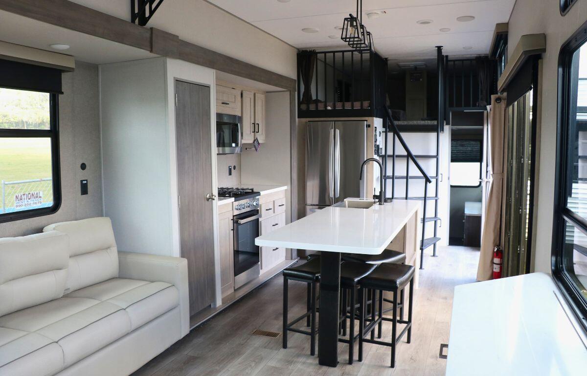 The interior of an RV features an open kitchen with a kitchen island, long sofa, cabinet, separate bedroom and stairs leading up to a sleeping area. Large windows light the inside of the RV.