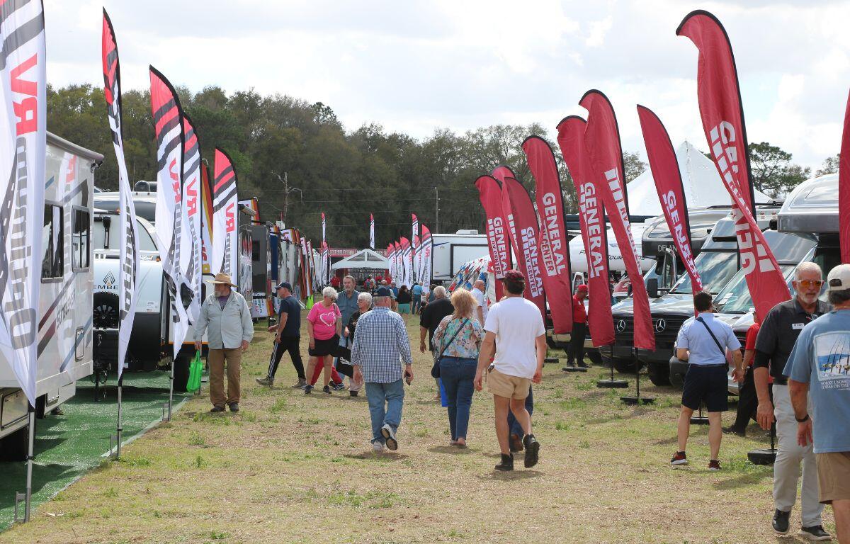 A grass field features RVs and mobile homes lined up with a large, open center where guests chat and walk around. Large flags reading, "Optimum RV" and "General RV Center."