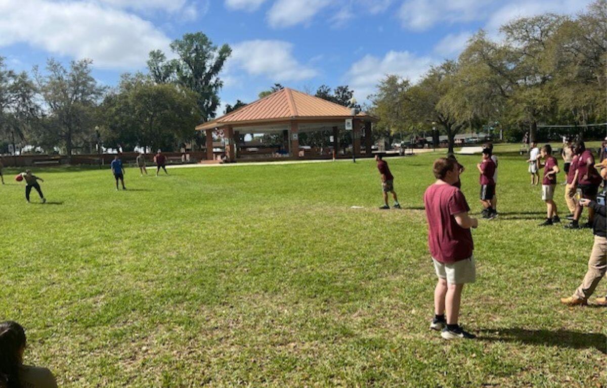 A bunch of young people in the background on a grass field, with trees and a large gazebo, in the foreground more people.