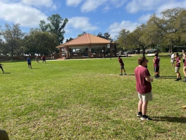 A bunch of young people in the background on a grass field, with trees and a large gazebo, in the foreground more people.