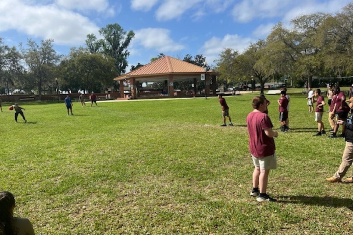 A bunch of young people in the background on a grass field, with trees and a large gazebo, in the foreground more people.