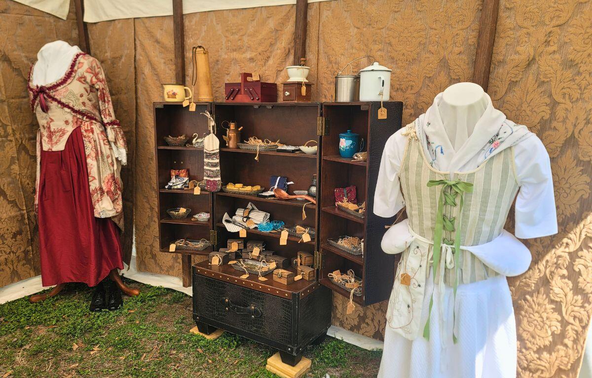 Historic garments are displayed on mannequins in a patch of grass inside of a canopy. Between the mannequins is a wardrobe with various historic items.