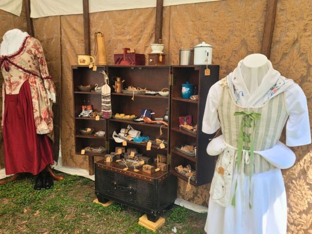 Historic garments are displayed on mannequins in a patch of grass inside of a canopy. Between the mannequins is a wardrobe with various historic items.