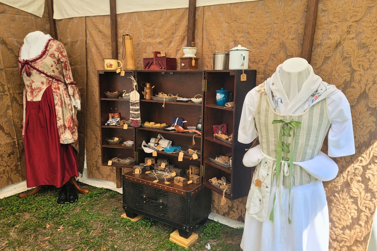 Historic garments are displayed on mannequins in a patch of grass inside of a canopy. Between the mannequins is a wardrobe with various historic items.