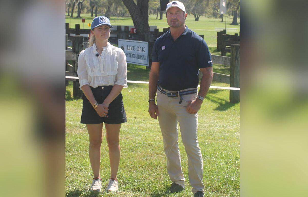 A woman on the left wearing a ball cap, white blouse and blue skirt standing next to a man wearing a white ball cap, blue shirt and tan pants, standing on grass, in front of a marathon course combined driving hazard with a placard with an insignia and writing attached to the hazard, with live oak trees and Spanish moss hanging in the background.