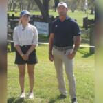 A woman on the left wearing a ball cap, white blouse and blue skirt standing next to a man wearing a white ball cap, blue shirt and tan pants, standing on grass, in front of a marathon course combined driving hazard with a placard with an insignia and writing attached to the hazard, with live oak trees and Spanish moss hanging in the background.
