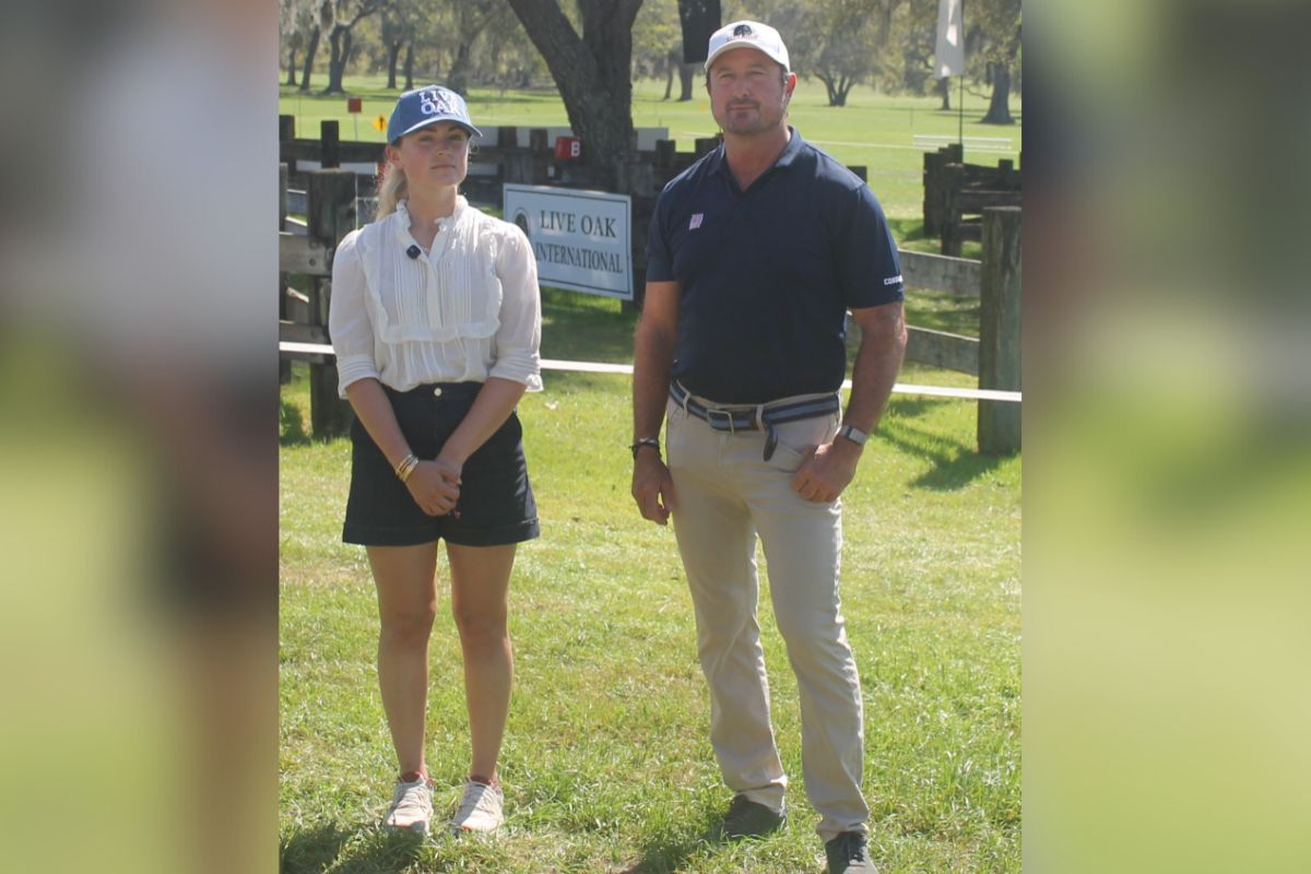 A woman on the left wearing a ball cap, white blouse and blue skirt standing next to a man wearing a white ball cap, blue shirt and tan pants, standing on grass, in front of a marathon course combined driving hazard with a placard with an insignia and writing attached to the hazard, with live oak trees and Spanish moss hanging in the background.