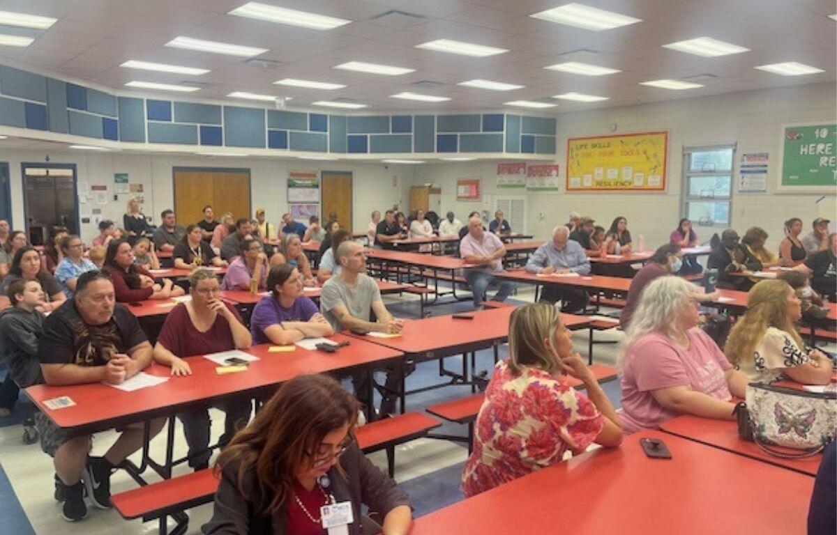 People sitting at tables, in a school cafeteria, with overhead lights, pictures and mosaics on the wall, with the audience fixated on the speaker, just off to the far right.