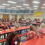People sitting at tables, in a school cafeteria, with overhead lights, pictures and mosaics on the wall, with the audience fixated on the speaker, just off to the far right.