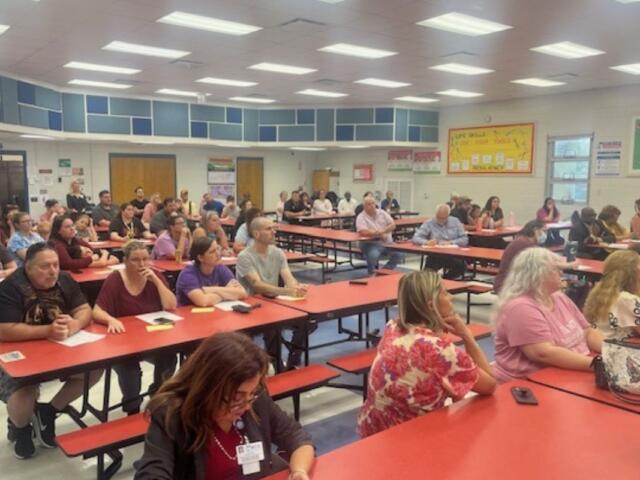 People sitting at tables, in a school cafeteria, with overhead lights, pictures and mosaics on the wall, with the audience fixated on the speaker, just off to the far right.