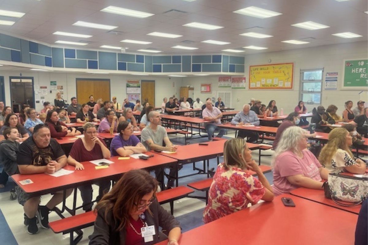 People sitting at tables, in a school cafeteria, with overhead lights, pictures and mosaics on the wall, with the audience fixated on the speaker, just off to the far right.