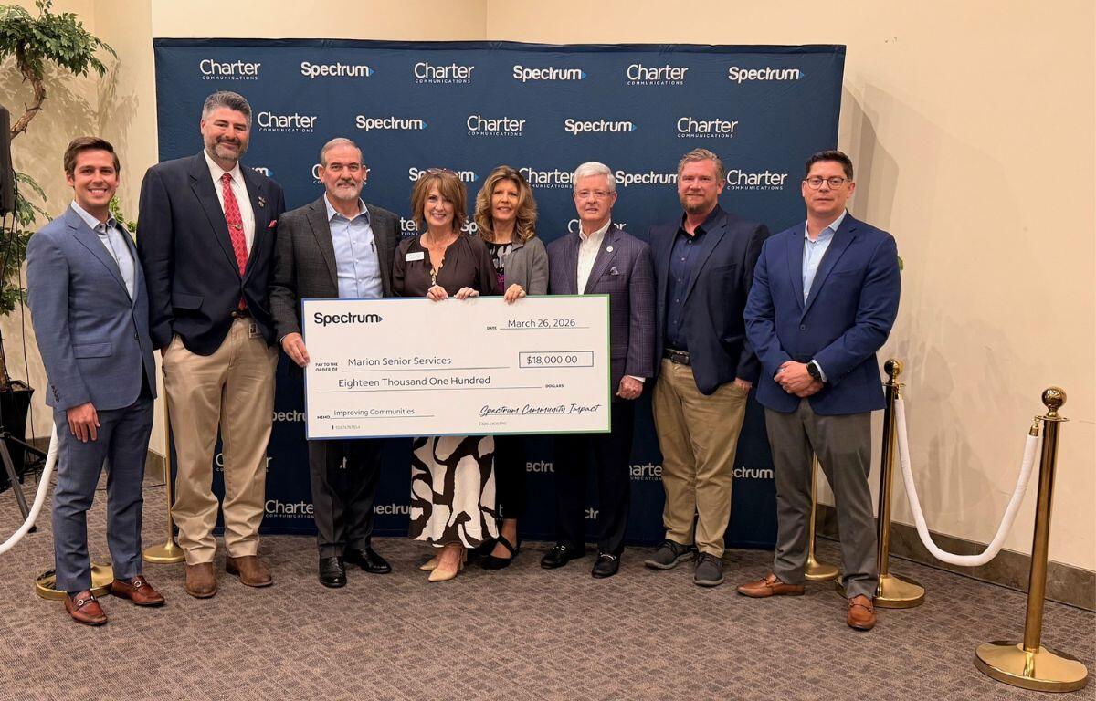 Brass stand to the left, people standing holding a large check in front of a blue banner, with a brass stand to the right.