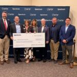 Brass stand to the left, people standing holding a large check in front of a blue banner, with a brass stand to the right.