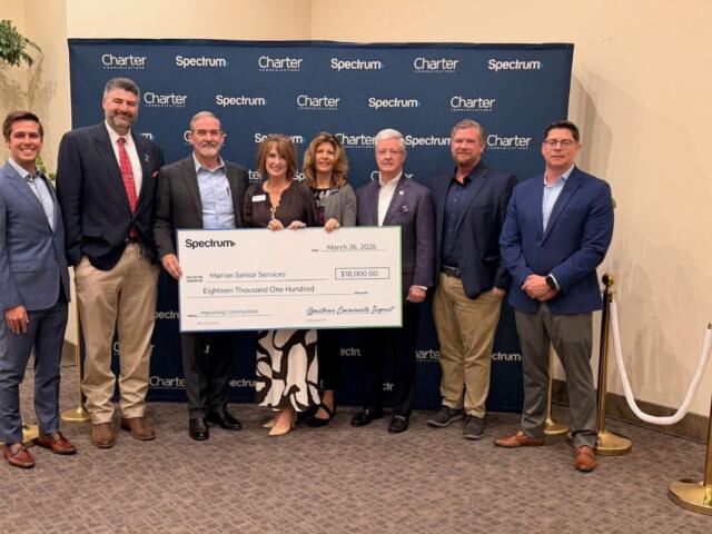 Brass stand to the left, people standing holding a large check in front of a blue banner, with a brass stand to the right.