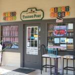 The exterior of a candy store features a gray door with two large windows on either side of the door with gray borders. In front of the window on the right is a tall table with three tall seats around it. A palm plant pokes into view in front of the shop. Above the door is a green, wooden sign that reads, "Wilderness Trading Post." On either side of the sign are colorful signs reading, "Sweet Janes Candy Store."
