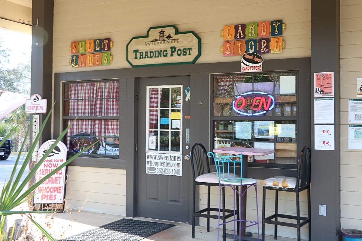 The exterior of a candy store features a gray door with two large windows on either side of the door with gray borders. In front of the window on the right is a tall table with three tall seats around it. A palm plant pokes into view in front of the shop. Above the door is a green, wooden sign that reads, "Wilderness Trading Post." On either side of the sign are colorful signs reading, "Sweet Janes Candy Store."