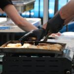 A man makes grilled cheese on a small burner while a person behind him prepared a carrier for the food.