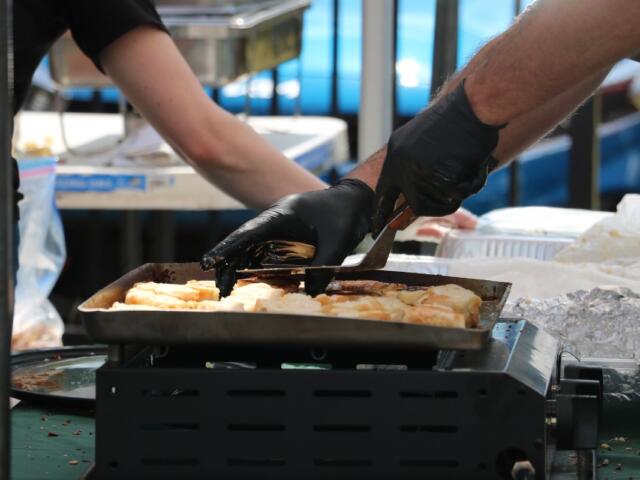 A man makes grilled cheese on a small burner while a person behind him prepared a carrier for the food.